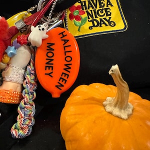May include: An orange pumpkin with a beige stem sits next to a keychain with a white ghost charm and an orange Halloween-themed tag that reads "HALLOWEEN MONEY". A black hoodie with a "HAVE A NICE DAY" patch is in the background.