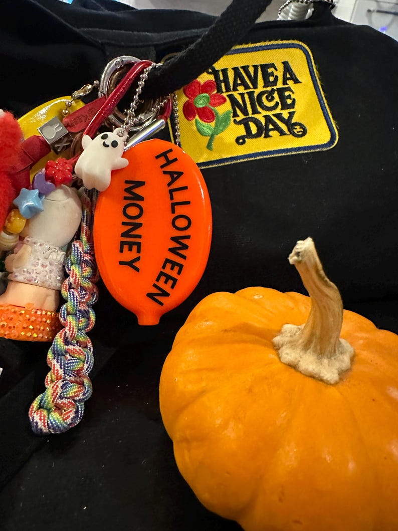 May include: An orange pumpkin with a beige stem sits next to a keychain with a white ghost charm and an orange Halloween-themed tag that reads "HALLOWEEN MONEY". A black hoodie with a "HAVE A NICE DAY" patch is in the background.