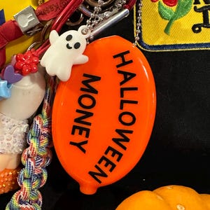 May include: An orange Halloween-themed charm with the words "HALLOWEEN MONEY" in black. A small white ghost charm is attached. The charm is part of a collection of keychains and accessories.