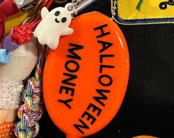 May include: An orange Halloween-themed charm with the words "HALLOWEEN MONEY" in black. A small white ghost charm is attached. The charm is part of a collection of keychains and accessories.