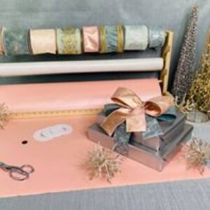 Puede incluir: Un juego de envoltura de regalos con papel de regalo rosa, un soporte de madera para envolver regalos con rollos de cinta de varios colores, incluyendo dorado, rosa y azul, y una pila de regalos envueltos con una cinta rosa y dorada.