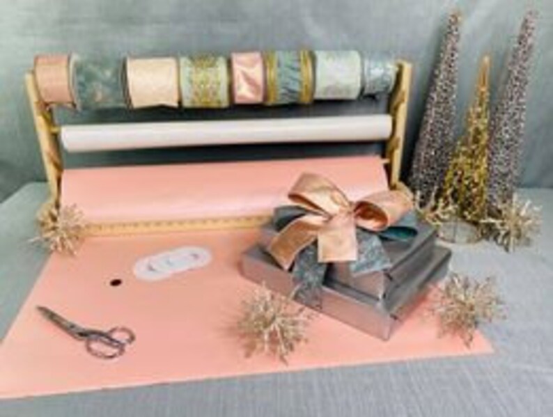 Puede incluir: Un juego de envoltura de regalos con papel de regalo rosa, un soporte de madera para envolver regalos con rollos de cinta de varios colores, incluyendo dorado, rosa y azul, y una pila de regalos envueltos con una cinta rosa y dorada.