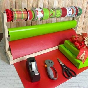 Puede incluir: Un soporte de papel de regalo de madera con dos rollos de papel de regalo, uno rojo y otro verde. El soporte tambi&eacute;n tiene un estante para guardar cintas y otros suministros de envoltura. Hay un dispensador de cinta, un par de tijeras y un cortador de papel en la mesa.