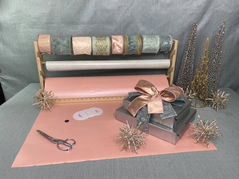 Puede incluir: Un rollo de papel de regalo rosa con un soporte de madera que sostiene rollos de cinta en varios tonos de rosa y verde. El soporte tiene una regla en la parte inferior. Hay dos cajas de regalo envueltas en papel azul y verde con un lazo rosa en la parte superior. Tambi&eacute;n hay estrellas doradas decorativas en la mesa.