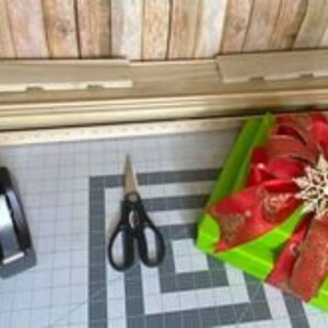 Puede incluir: Una caja de regalo verde con un lazo rojo y un adorno de copo de nieve dorado. La caja est&aacute; sobre una mesa con un dispensador de cinta adhesiva, tijeras y una herramienta de envoltura de caja de regalo de madera.