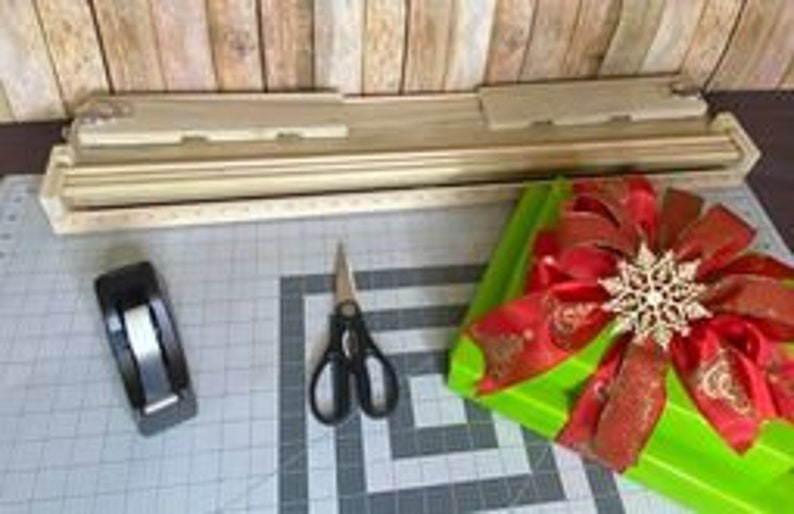Puede incluir: Una caja de regalo verde con un lazo rojo y un adorno de copo de nieve dorado. La caja est&aacute; sobre una mesa con un dispensador de cinta adhesiva, tijeras y una herramienta de envoltura de caja de regalo de madera.