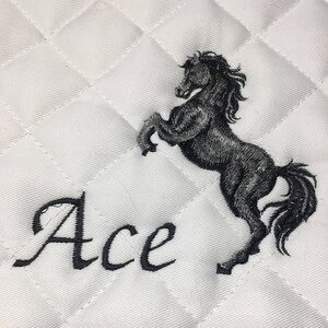 May include: White quilted saddle pad with a black embroidered horse and the word "Ace" in a cursive font.