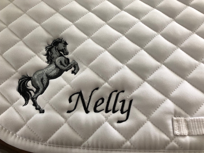 May include: White quilted saddle pad with a black embroidered horse and the name "Nelly" in cursive.