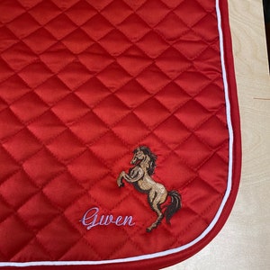 May include: Red quilted saddle pad with a white border. The pad features an embroidered horse and the name "Gwen".