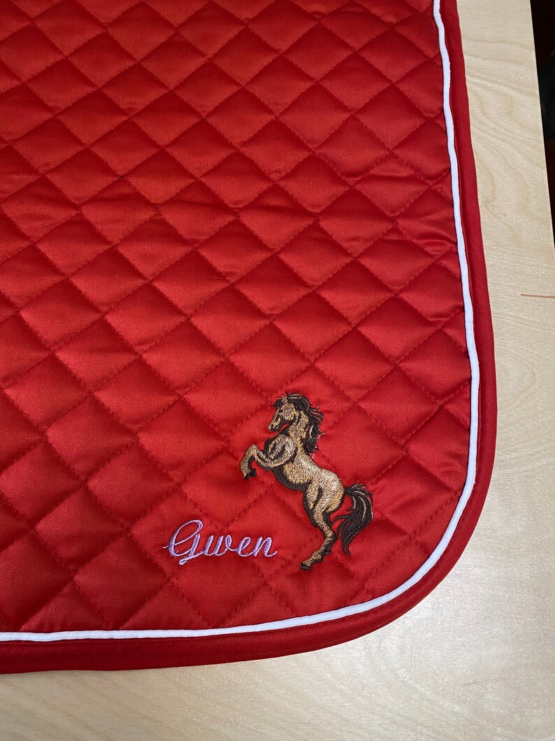 May include: Red quilted saddle pad with a white border. The pad features an embroidered horse and the name "Gwen".