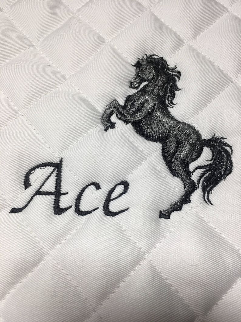May include: White quilted saddle pad with a black embroidered horse and the word "Ace" in a cursive font.