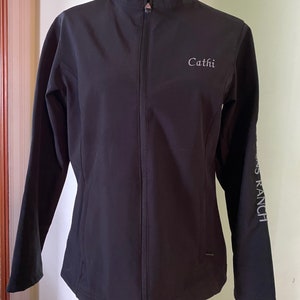 May include: Black softshell jacket with a zipper closure and a stand-up collar. The jacket has the name "Cathi" embroidered on the front and "S Ranch" on the sleeve.