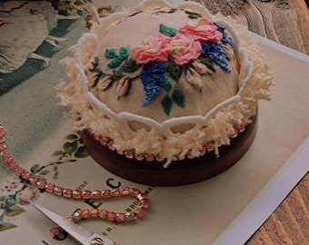 May include: A vintage-style pincushion with a floral embroidery design in pink, blue, and green. The pincushion sits on a wooden base, surrounded by lace trim. A pair of decorative scissors and a beaded chain are also present.