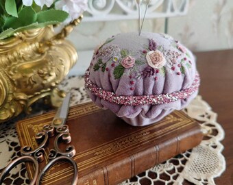 May include: A purple velvet pincushion with embroidered flowers and a pink beaded trim. Two white pins are stuck in the top. The pincushion is sitting on a brown book with gold lettering.
