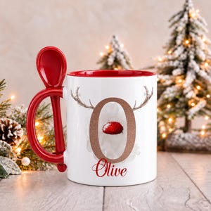 May include: A white ceramic mug with a red interior and a matching red spoon integrated into the handle. The front features a large letter O with reindeer antlers and a red nose, with the name Olive printed in red script below the letter.