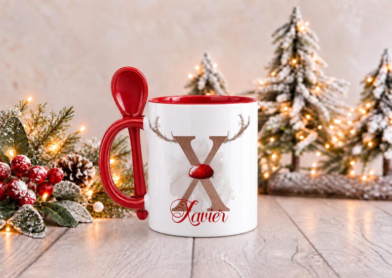May include: A white ceramic mug with a red interior and a matching red spoon integrated into the handle. The front features a large brown letter X with reindeer antlers and a red nose, with the name Xavier written in red script below.
