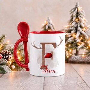 May include: A white ceramic mug with a red interior and a matching red spoon integrated into the handle. The front features a large brown letter F with reindeer antlers and a red nose, with the name Finn printed in red script below.