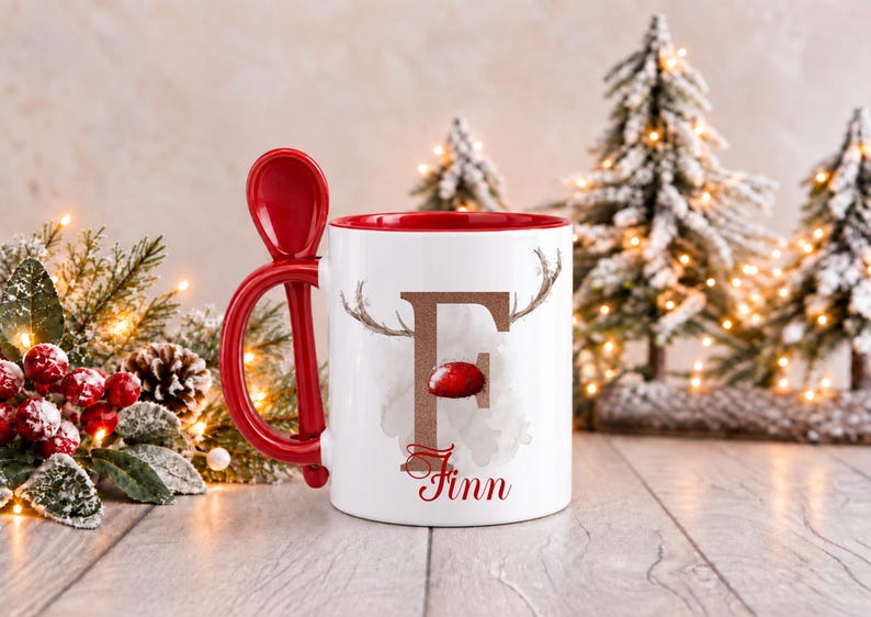 May include: A white ceramic mug with a red interior and a matching red spoon integrated into the handle. The front features a large brown letter F with reindeer antlers and a red nose, with the name Finn printed in red script below.