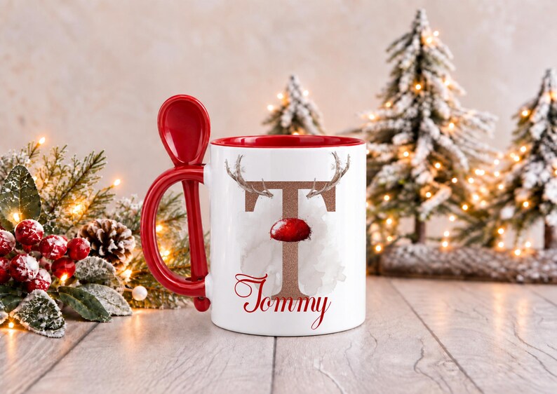May include: A white ceramic mug with a red interior and a matching red spoon integrated into the handle. The front features a large brown letter T decorated with reindeer antlers and a red nose, with the name Tommy written in red script below.