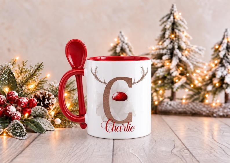 May include: A white ceramic mug with a red interior and a matching red spoon integrated into the handle. The front features a large brown letter C with reindeer antlers and a red nose, with the name Charlie written in red script below.