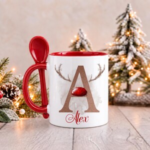 May include: A white ceramic mug with a red interior and a matching red spoon integrated into the handle. The front features a large brown letter A with reindeer antlers and a red nose, with the name Alex written in red script below.