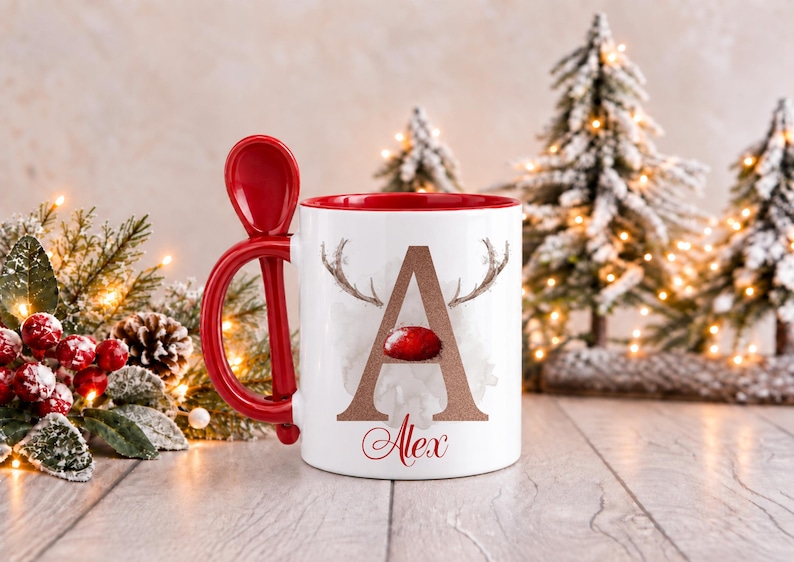 May include: A white ceramic mug with a red interior and a matching red spoon integrated into the handle. The front features a large brown letter A with reindeer antlers and a red nose, with the name Alex written in red script below.