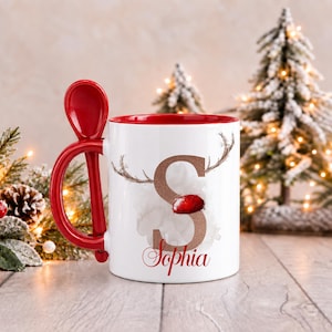May include: A white ceramic mug with a red interior and a matching red spoon integrated into the handle. The front features a large glittery bronze letter S with reindeer antlers and a red nose, with the name Sophia printed below in red.