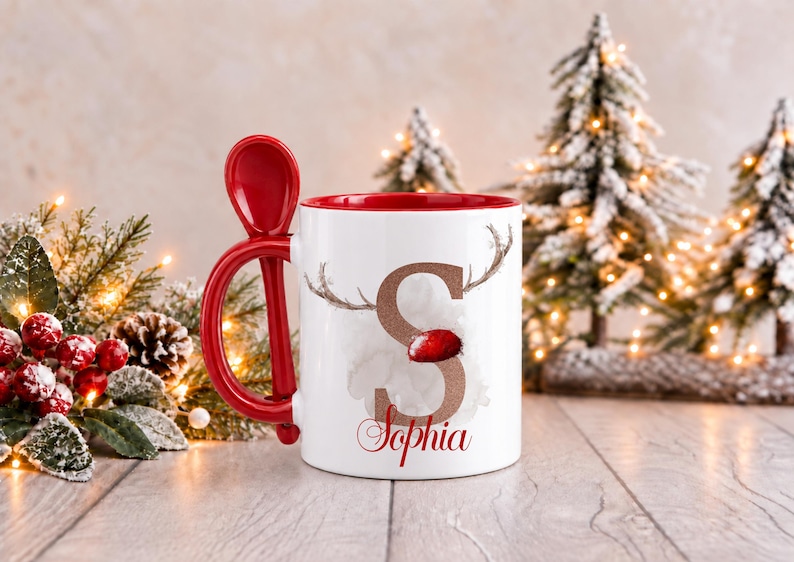 May include: A white ceramic mug with a red interior and a matching red spoon integrated into the handle. The front features a large glittery bronze letter S with reindeer antlers and a red nose, with the name Sophia printed below in red.