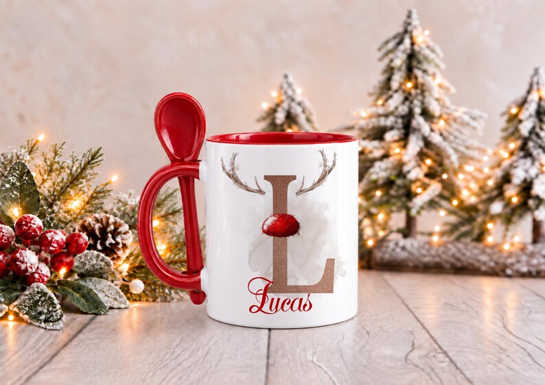 May include: A white ceramic mug with a red interior and handle, featuring a red spoon attached to the side. The front displays a large letter L with reindeer antlers and a red nose, with the name Lucas printed in red script underneath.