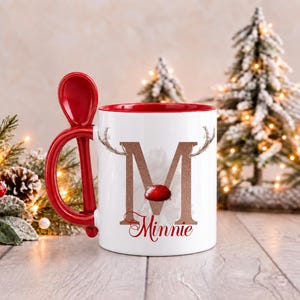 May include: A white ceramic mug with a red interior and a matching red spoon integrated into the handle. The front features a large glittery copper letter M adorned with reindeer antlers and a red nose, with the name Minnie printed below in red.