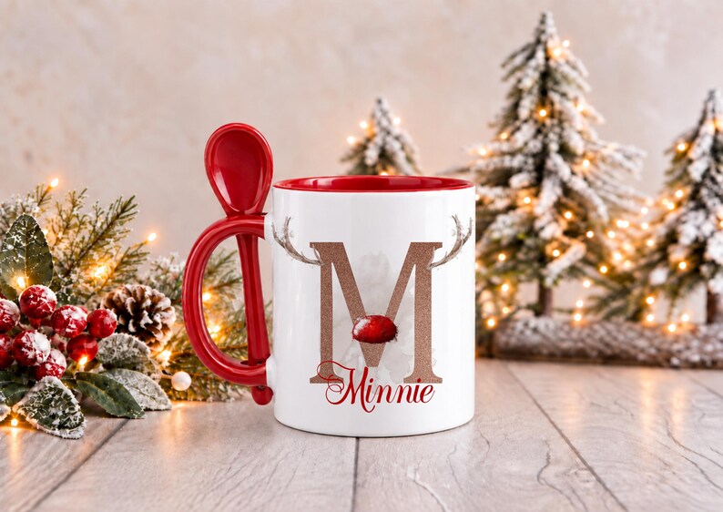 May include: A white ceramic mug with a red interior and a matching red spoon integrated into the handle. The front features a large glittery copper letter M adorned with reindeer antlers and a red nose, with the name Minnie printed below in red.