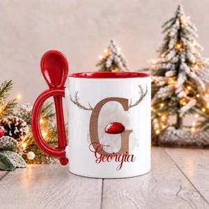 May include: A white ceramic mug with a red interior and a matching red spoon integrated into the handle. The mug features a large brown letter G with reindeer antlers and a red nose, with the name Georgia printed in red script below it.