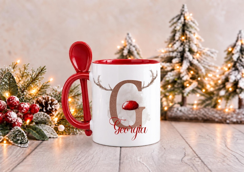 May include: A white ceramic mug with a red interior and a matching red spoon integrated into the handle. The mug features a large brown letter G with reindeer antlers and a red nose, with the name Georgia printed in red script below it.