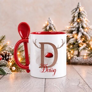 May include: A white ceramic mug with a red interior and a matching red spoon integrated into the handle. The front features a large letter D with reindeer antlers and a red nose, with the name Daisy printed in red script below it.