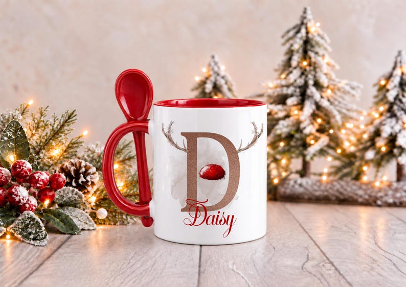 May include: A white ceramic mug with a red interior and a matching red spoon integrated into the handle. The front features a large letter D with reindeer antlers and a red nose, with the name Daisy printed in red script below it.