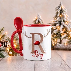 May include: A white ceramic mug with a red interior and a red handle featuring an integrated spoon slot. The exterior displays a large, glittery bronze letter R with reindeer antlers and a red nose, with the name Rory printed in red script below.