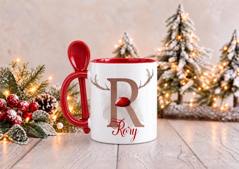 May include: A white ceramic mug with a red interior and a red handle featuring an integrated spoon slot. The exterior displays a large, glittery bronze letter R with reindeer antlers and a red nose, with the name Rory printed in red script below.