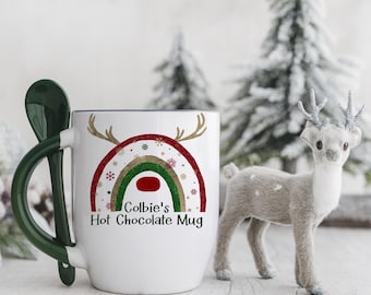 May include: A white ceramic mug with a dark green handle and an integrated spoon slot. The front features a rainbow graphic in red, green, and gold with reindeer antlers and snowflakes. The text reads Colbie's Hot Chocolate Mug in black.
