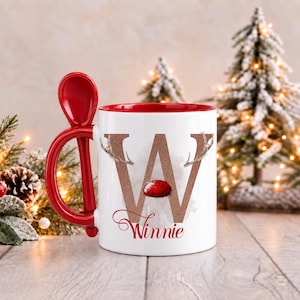May include: A white ceramic mug with a red interior and a matching red spoon integrated into the handle. The front features a large, glittery copper letter W decorated with reindeer antlers and a red nose, with the name Winnie written below.