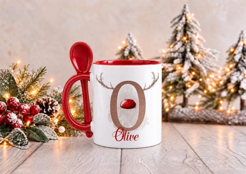 May include: A white ceramic mug with a red interior and a matching red spoon integrated into the handle. The front features a large letter O with reindeer antlers and a red nose, with the name Olive printed in red script below the letter.