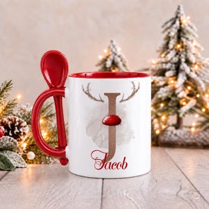 May include: A white ceramic mug with a red interior and a matching red spoon integrated into the handle. The front features a large letter J with reindeer antlers and a red nose, with the name Jacob printed in red script below it.