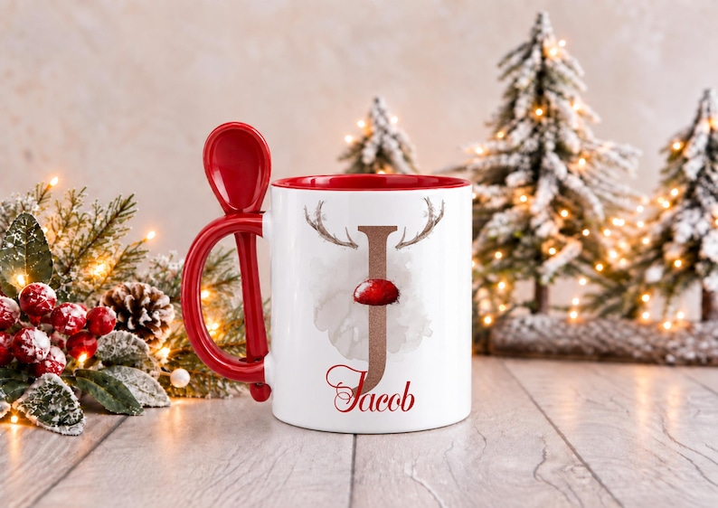 May include: A white ceramic mug with a red interior and a matching red spoon integrated into the handle. The front features a large letter J with reindeer antlers and a red nose, with the name Jacob printed in red script below it.