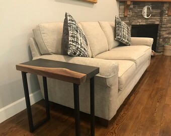 May include: A modern side table with a live edge walnut top and a black epoxy resin inlay. The table has black metal legs. The table is placed next to a beige sofa with patterned pillows. The room has hardwood floors.