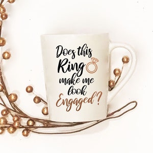 May include: White ceramic mug with black text that reads "Does this Ring make me look Engaged?" with a gold ring graphic.