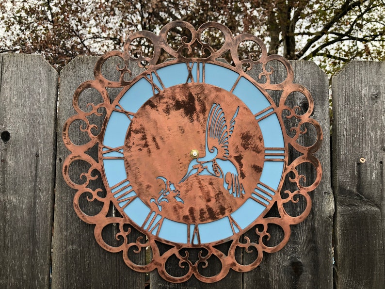 May include: A copper-colored metal clock with a blue face and a hummingbird design. The clock is surrounded by a decorative metal frame.