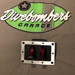 Peut inclure: Un panneau métallique avec deux interrupteurs rouges éclairés. Le panneau est monté sur une surface grise, avec le texte "Divebombers Garage" au-dessus. Le texte est en blanc et vert, avec un logo d'étoile.