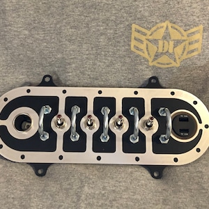 Puede incluir: Un panel de metal plateado con seis interruptores de palanca y un puerto USB doble. El panel tiene un fondo negro y está rodeado por un borde plateado. Los interruptores son rojos y negros.