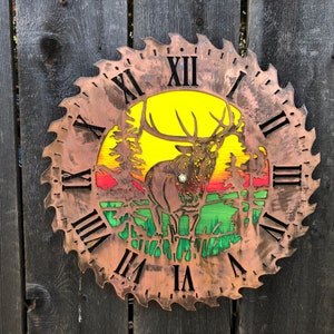 May include: A rustic clock made from a repurposed saw blade with a colorful elk silhouette and a sunset scene. The clock face features Roman numerals and a copper-toned finish.