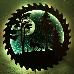 Op de afbeelding: Een cirkelvormig metalen wandkunstwerk met een silhouet van een wolf die huilt naar de maan in een bosrijke omgeving. De maan is verlicht met een groene gloed, en de achtergrond is een sterrenhemel.