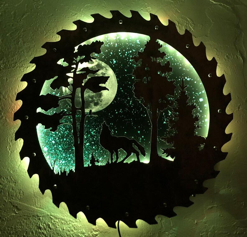 Op de afbeelding: Een cirkelvormig metalen wandkunstwerk met een silhouet van een wolf die huilt naar de maan in een bosrijke omgeving. De maan is verlicht met een groene gloed, en de achtergrond is een sterrenhemel.